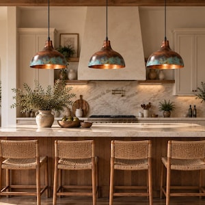 May include: Three dome-shaped pendant lights with a weathered copper and teal patina finish hang above a kitchen island. The lights feature a smooth, rounded exterior and a warm metallic interior, suspended by thin black cords from the ceiling.