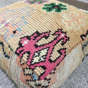 May include: A colorful, hand-woven pouf with a pink, black, and white geometric pattern. The pouf is made of a soft, plush fabric and has a square shape.