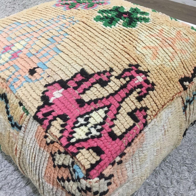 May include: A colorful, hand-woven pouf with a pink, black, and white geometric pattern. The pouf is made of a soft, plush fabric and has a square shape.