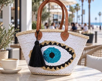 Puede incluir: Un bolso tipo tote de paja tejida blanca con un diseño grande de ojo turco hecho de lentejuelas en azul, negro y dorado. El bolso tiene dos asas de cuero marrón sujetas con pestañas cosidas y una borla negra larga colgando de una base.