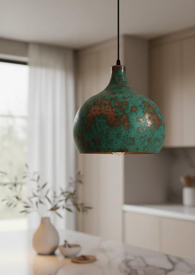 Puede incluir: L&aacute;mpara colgante con acabado verde y cobre. La l&aacute;mpara tiene una pantalla redondeada en forma de campana con una superficie texturizada. La luz cuelga de un cable negro, en un entorno de cocina.