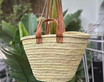 May include: A woven straw tote bag with brown leather handles. The bag is hanging from a tree branch.