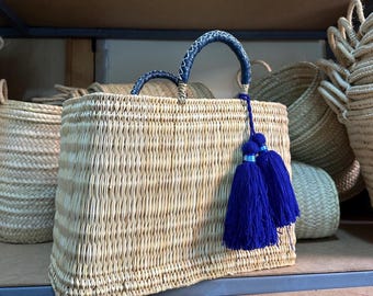 May include: Woven straw tote bag with blue handles and tassel accents. The rectangular bag features a natural beige color with a woven pattern. The handles are a dark blue with white stitching. Two blue tassels hang from the side.