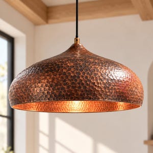 May include: A dome-shaped pendant light fixture featuring a hammered copper exterior finish and a smooth, reflective interior surface. The lamp is suspended from a thin black cord attached to a small brass-colored metal fitting at the top.