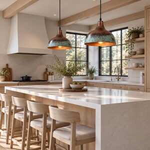 May include: Two dome-shaped pendant lights with a weathered copper and teal patina finish hang above a large kitchen island. The island features a white stone countertop and light wood cabinetry, paired with four wooden stools with beige cushions.