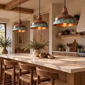 May include: Three dome-shaped pendant lights with a weathered copper and teal patina finish hang above a kitchen island. The lights feature a smooth, rounded silhouette with a metallic interior that reflects the warm glow of the light bulbs.