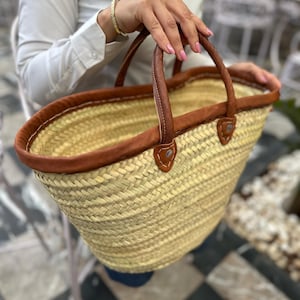May include: A large, woven straw tote bag with brown leather handles and trim. The bag has a wide, oval shape and is perfect for carrying groceries or other items.