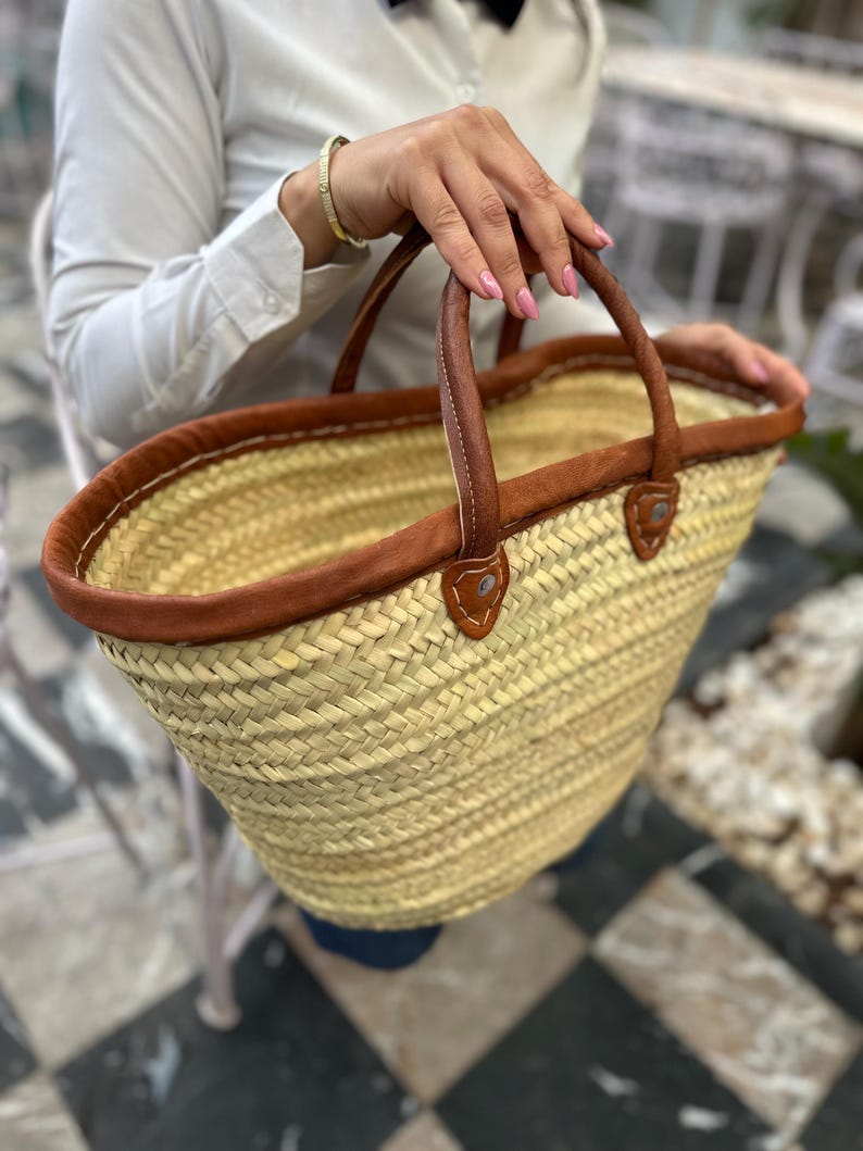 May include: A large, woven straw tote bag with brown leather handles and trim. The bag has a wide, oval shape and is perfect for carrying groceries or other items.