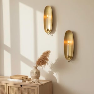 May include: Two gold wall sconces with lit candles. Each sconce has an oval backplate and a curved holder for the candle. A vase with dried plants and a wooden cabinet are in the foreground.