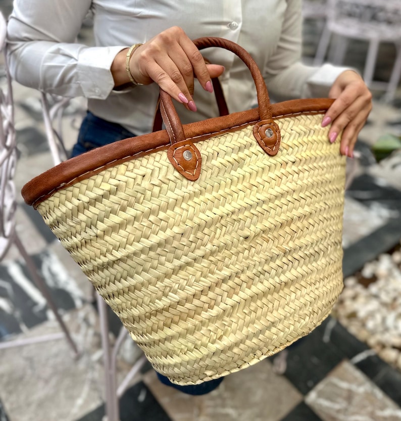 May include: A large, woven straw tote bag with brown leather trim and handles. The bag has a wide, open top and a rounded bottom.
