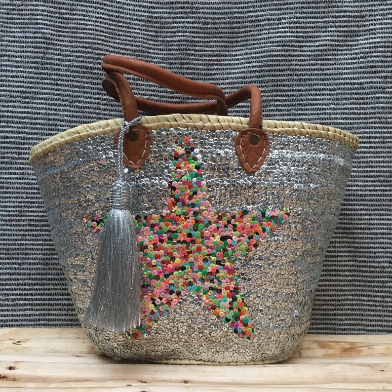 May include: A silver sequined tote bag with a star design made of colorful sequins. The bag has brown leather handles and a silver tassel. The bag is made of woven straw.