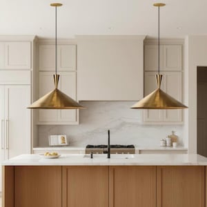 May include: Two brass pendant lights hang above a kitchen island. Each light has a conical shade and a slender black cord. The kitchen features light wood cabinetry, a white countertop, and a marble backsplash.