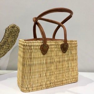 Op de afbeelding: Een geweven strooien tas met bruine leren handvatten en een bruine leren bies. De tas heeft een rechthoekige vorm en is gemaakt van natuurlijke materialen.