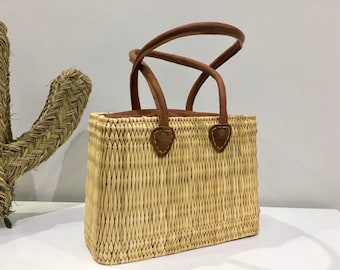 May include: A woven straw tote bag with brown leather handles and a brown leather trim. The bag has a rectangular shape and is made of natural materials.