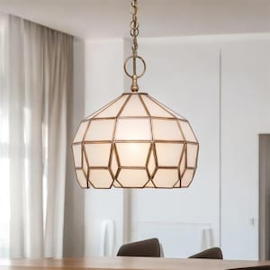 Puede incluir: Una lámpara colgante geométrica con una pantalla de vidrio blanco y un marco de metal color bronce. La luz cuelga de una cadena y está suspendida sobre una mesa de madera. El accesorio tiene un diseño único y multifacético.