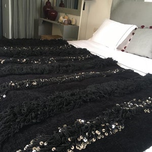 Op de afbeelding: Zwarte deken met een franje rand en zilveren pailletten. De deken ligt op een wit bed met een grijs hoofdeinde.