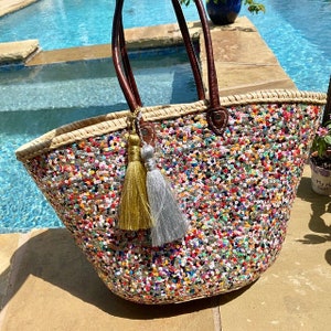 May include: A large straw tote bag with a colourful sequin design. The bag has two brown leather handles and two tassel charms, one gold and one silver.