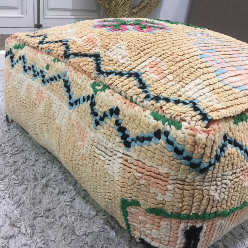 May include: A square, beige pouf with a colorful geometric pattern. The pouf is made of woven fabric and has a fringed edge.