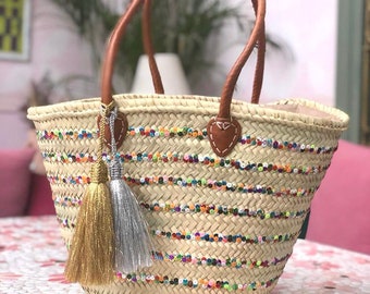 Puede incluir: Un bolso de mimbre con asas de cuero y ribete de cuentas de colores. El bolso tiene dos borlas, una dorada y otra plateada.
