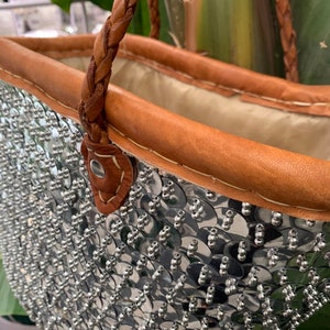 K&ouml;nnte beinhalten: Eine silberne Pailleten-Tragetasche mit braunem Lederbesatz und geflochtenen Griffen. Die Au&szlig;enseite der Tasche ist mit gl&auml;nzenden, runden Pailletten bedeckt, die eine strukturierte, reflektierende Oberfl&auml;che bilden. Das Innenfutter ist hellbeige.