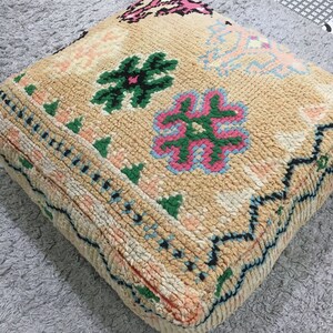 May include: A beige floor cushion with a colorful floral pattern. The cushion has a black border and is made of a soft, woven fabric.