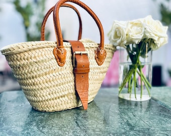 May include: A woven straw tote bag with brown leather handles and a buckle closure. The bag is sitting on a green marble surface.