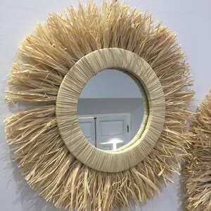 May include: A round mirror with a natural woven straw frame. The frame has a thick, fringed edge. The mirror is hanging on a white wall.
