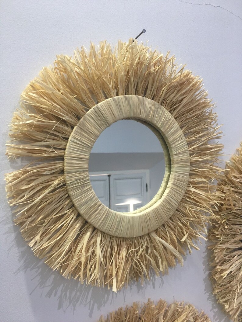 May include: A round mirror with a natural woven straw frame. The frame has a thick, fringed edge. The mirror is hanging on a white wall.