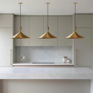 Puede incluir: Tres lámparas colgantes de latón con una pantalla cónica cuelgan sobre una encimera de mármol blanco. La cocina cuenta con gabinetes de color gris claro y una placa para salpicaduras de mármol. Las luces están suspendidas del techo por cables negros.