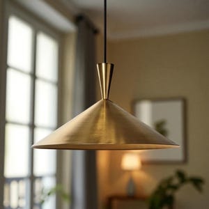 May include: A brass-coloured, cone-shaped pendant light fitting. The light has a wide, downward-facing shade and a slender, conical top. The fitting hangs from a black cord, and the background includes a window and a wall.