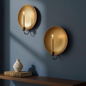 May include: Two gold wall sconces with lit candles. Each sconce has a hammered texture and a curved holder for the candle. A small white vase and a book sit on a wooden shelf below.