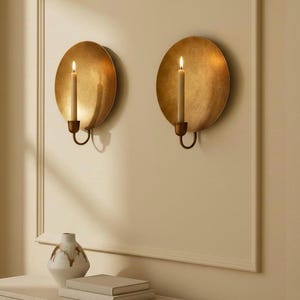 May include: Two antique brass wall sconces with lit candles. Each sconce features a circular backplate and a curved candle holder. The sconces are mounted on a cream-coloured wall, creating a warm and inviting ambience. The candles are approximately 15 cm tall.