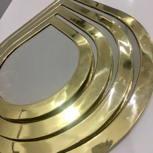 May include: A gold-toned, layered, decorative mirror with a teardrop shape. The mirror has a polished, reflective surface.
