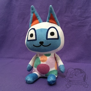 May include: A plush toy cat with a white face, blue eyes, and ears with orange accents. The cat's body is white with colorful polka dots. The toy is sitting on a purple surface.