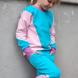 Puede incluir: Conjunto de dos piezas para niños con diseño de bloques de color. La parte superior de manga larga es rosa y turquesa, con ribete morado en el cuello y los puños. Los pantalones a juego son turquesa con detalles rosas. El niño está apoyado en una pared gris.