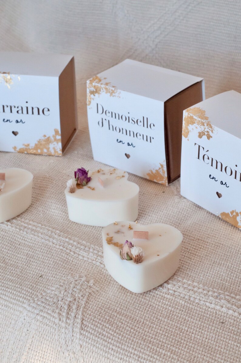 Op de afbeelding: Hartvormige witte kaarsen met gedroogde bloemen en gouden accenten. De kaarsen worden getoond met witte dozen met goudfoliedetails en Franse tekst. Een romantisch en elegant thema.