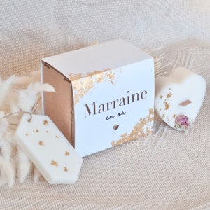 Op de afbeelding: Een witte geschenkdoos met goudfolie accenten en de tekst "Marraine en or". Een hartvormige kaars en een tag-vormige wax melt, beide met goudfolie. De items zijn gerangschikt op een neutrale stof.