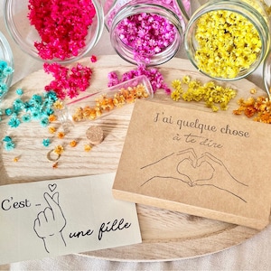 May include: A brown card with the text "J'ai quelque chose &agrave; te dire" and a drawing of two hands making a heart shape.  The card is surrounded by small dried flowers in various colors, including blue, pink, yellow, and orange.
