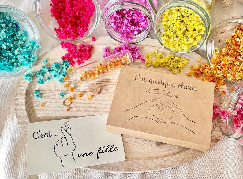 May include: A brown card with the text "J'ai quelque chose &agrave; te dire" and a drawing of two hands making a heart shape.  The card is surrounded by small dried flowers in various colors, including blue, pink, yellow, and orange.