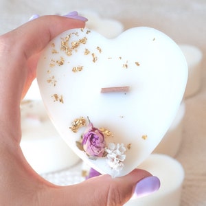 Op de afbeelding: Een witte hartvormige kaars met goudbladaccenten, een houten lont en gedroogde bloemen. De kaars heeft de tekst "Parfum Vanille - Figue". Andere hartvormige kaarsen zijn op de achtergrond te zien.