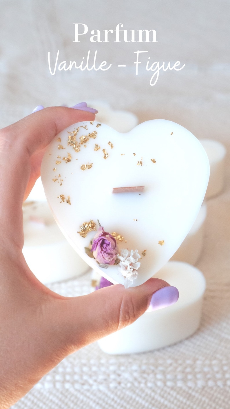 Op de afbeelding: Een witte hartvormige kaars met goudbladaccenten, een houten lont en gedroogde bloemen. De kaars heeft de tekst "Parfum Vanille - Figue". Andere hartvormige kaarsen zijn op de achtergrond te zien.
