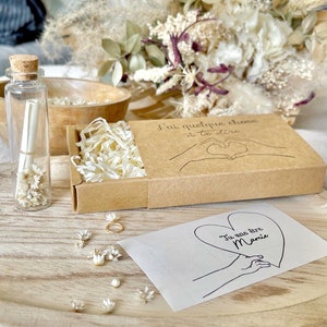 May include: A small brown box with a white paper insert and a handwritten message that says "J'ai quelque chose a te dire". The box is open and there is a small white envelope with a handwritten message that says "Tu vas &ecirc;tre Mamie" on it. There are also small white flowers scattered around the box.