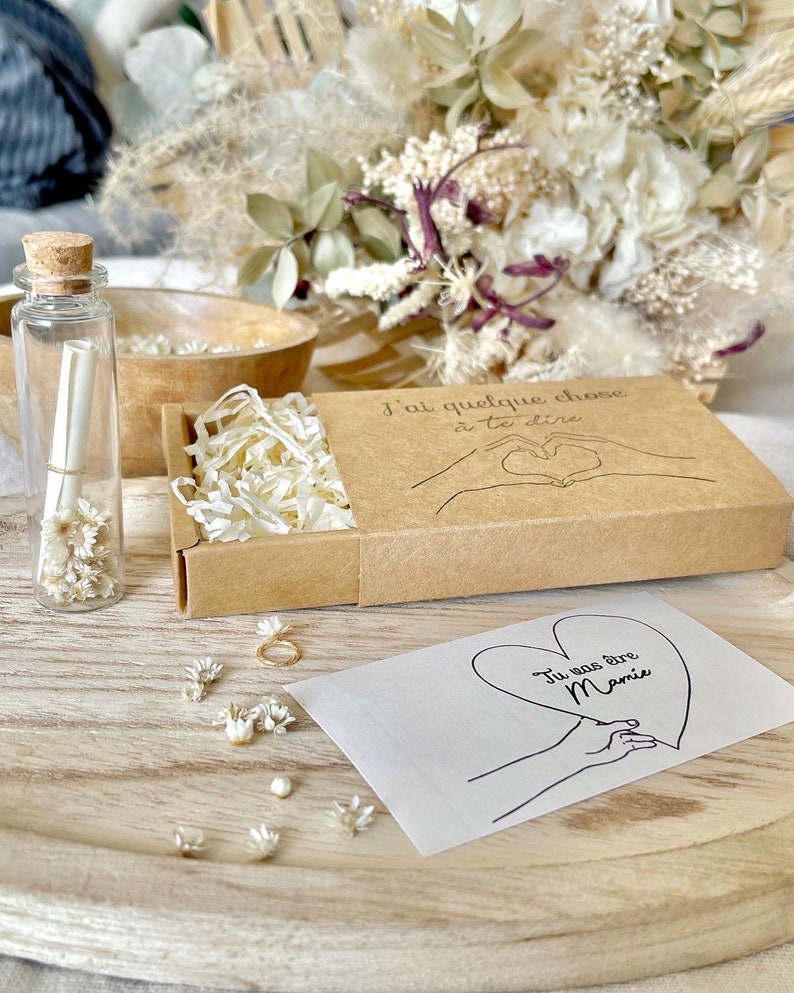 May include: A small brown box with a white paper insert and a handwritten message that says "J'ai quelque chose a te dire". The box is open and there is a small white envelope with a handwritten message that says "Tu vas &ecirc;tre Mamie" on it. There are also small white flowers scattered around the box.