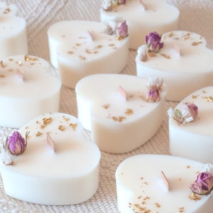 Op de afbeelding: Hartvormige witte kaarsen met houten lonten, versierd met gedroogde bloemen en goudvlokken. Het woord "Bougie" staat bovenaan. De kaarsen zijn gerangschikt op een witte, getextureerde ondergrond.