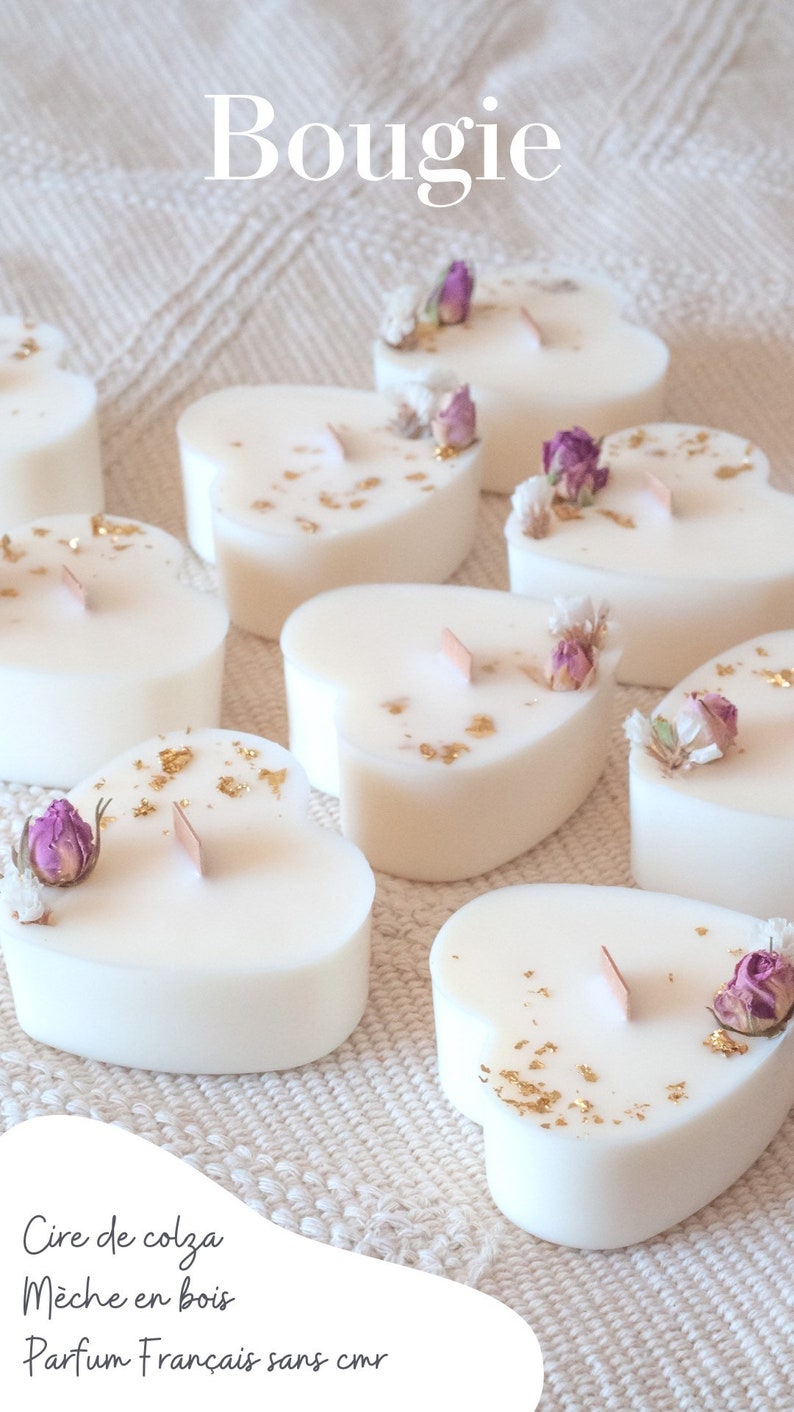 Op de afbeelding: Hartvormige witte kaarsen met houten lonten, versierd met gedroogde bloemen en goudvlokken. Het woord "Bougie" staat bovenaan. De kaarsen zijn gerangschikt op een witte, getextureerde ondergrond.