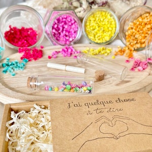 May include: A small brown box with a white paper filler inside. The box has a lid with the text "J'ai quelque chose &agrave; te dire" and a drawing of two hands making a heart shape. The box is surrounded by small glass jars filled with colorful dried flowers.