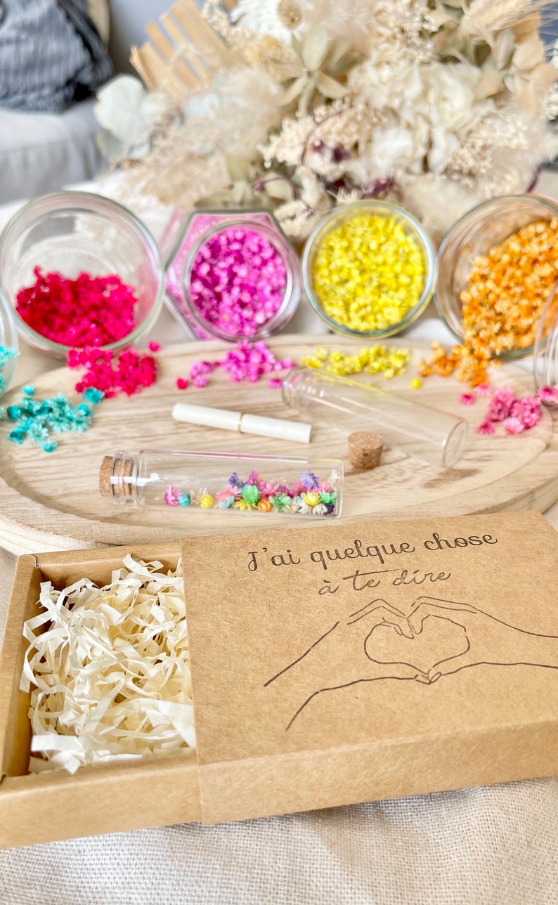 May include: A small brown box with a white paper filler inside. The box has a lid with the text "J'ai quelque chose &agrave; te dire" and a drawing of two hands making a heart shape. The box is surrounded by small glass jars filled with colorful dried flowers.
