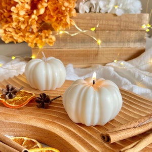 Op de afbeelding: Twee witte, pompoenvormige kaarsen op een houten snijplank met gedroogde sinaasappelschijfjes en kaneelstokjes. De kaarsen zijn aangestoken en hebben een geribbelde textuur.