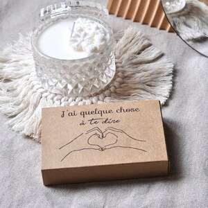 May include: A brown cardboard box with the text "J'ai quelque chose &agrave; te dire" and a hand drawn image of two hands making a heart shape. The box is sitting on a white textured surface.