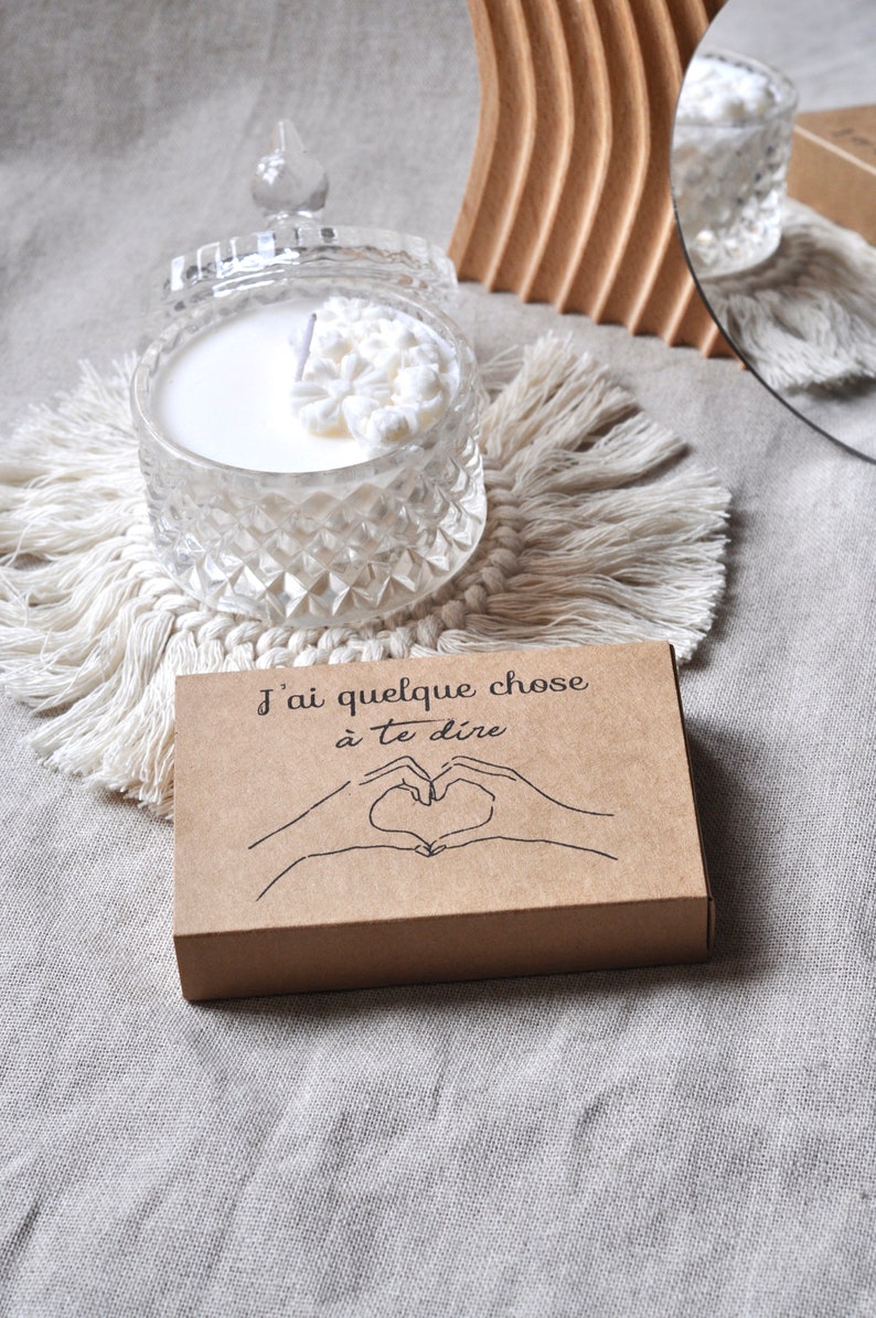 May include: A brown cardboard box with the text "J'ai quelque chose &agrave; te dire" and a hand drawn image of two hands making a heart shape. The box is sitting on a white textured surface.